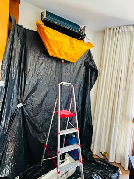 Higienização em andamento: parede e piso cobertos por lona preta, bolsa coletora amarela presa sob a unidade aberta e escada posicionada no centro do cômodo.